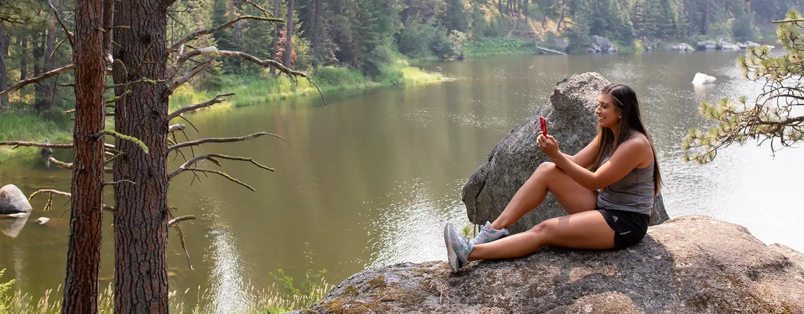 Woman in woods sitting by creek on smart phone smiling