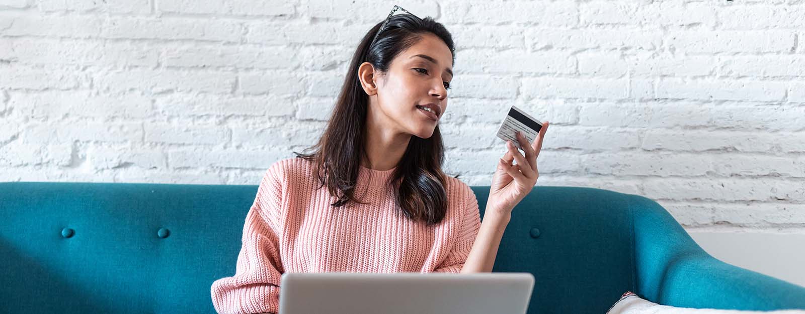 Woman on couch holding debit card while working on laptop
