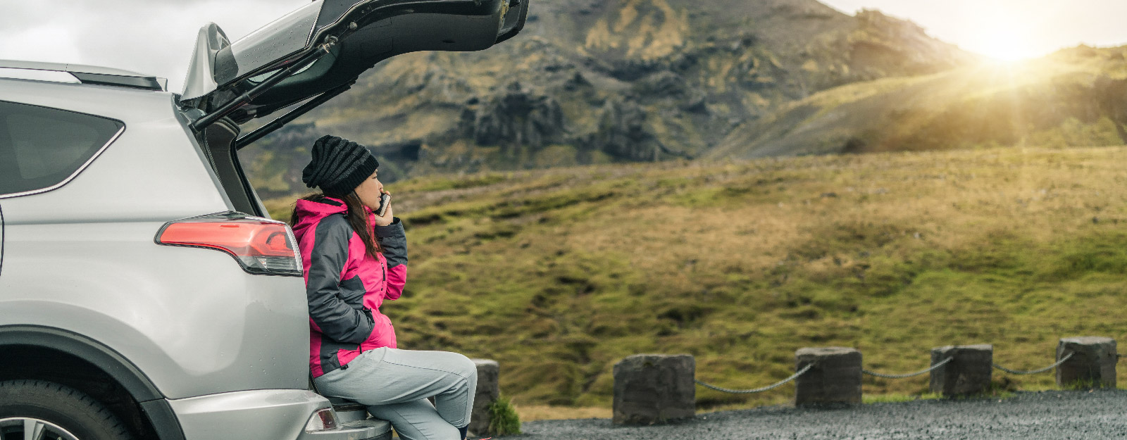 Person in hiking clothes on the side of the road sitting in their SUV trunk while on phone
