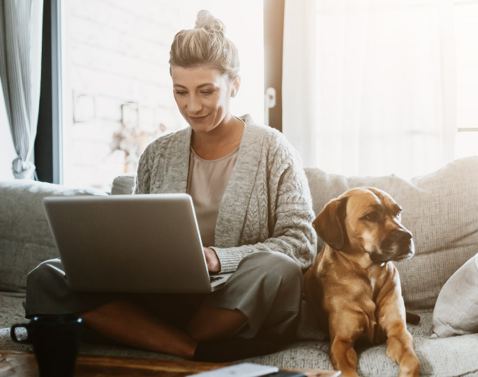 Woman on a computer while her dog sits on the couch net to her.